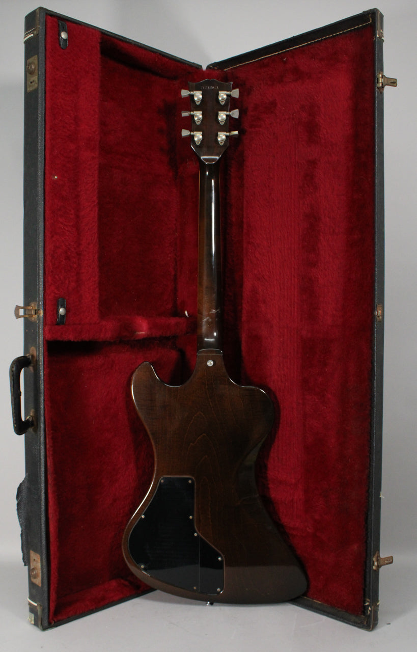 The dark walnut guitar, with its back facing the viewer, rests in a red velvet-lined case, showing some wear on its neck.