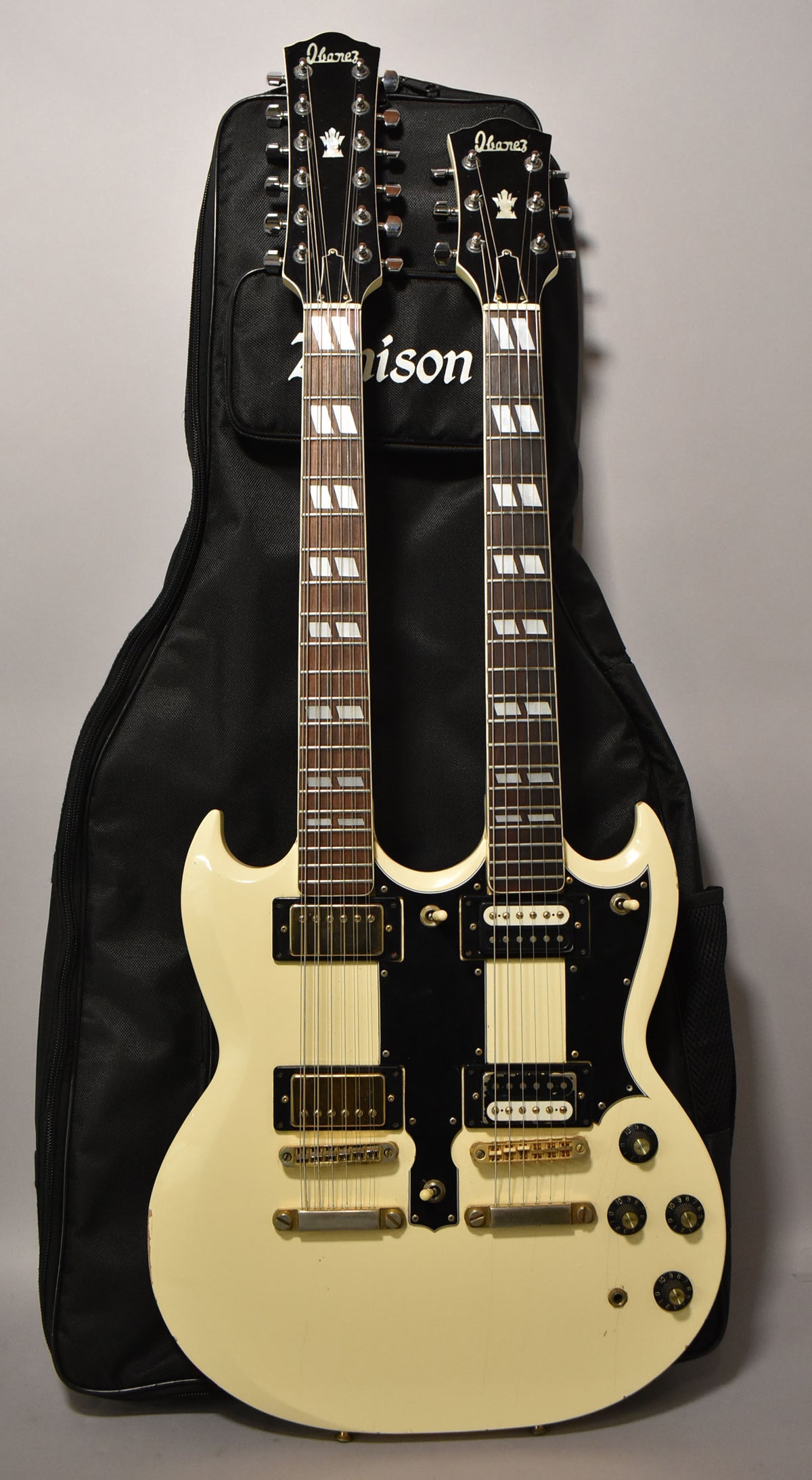 Full-length view of a cream double-neck electric guitar with a black pickguard, gold hardware, and a black gig bag behind it.