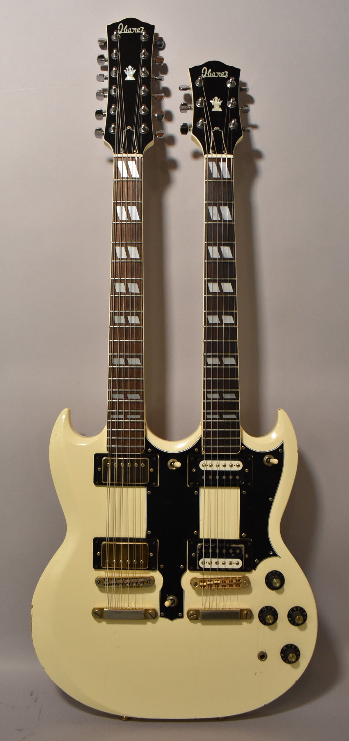 Full front view of a cream double-neck guitar with black pickguards, gold hardware, and block inlays, showing some finish wear.