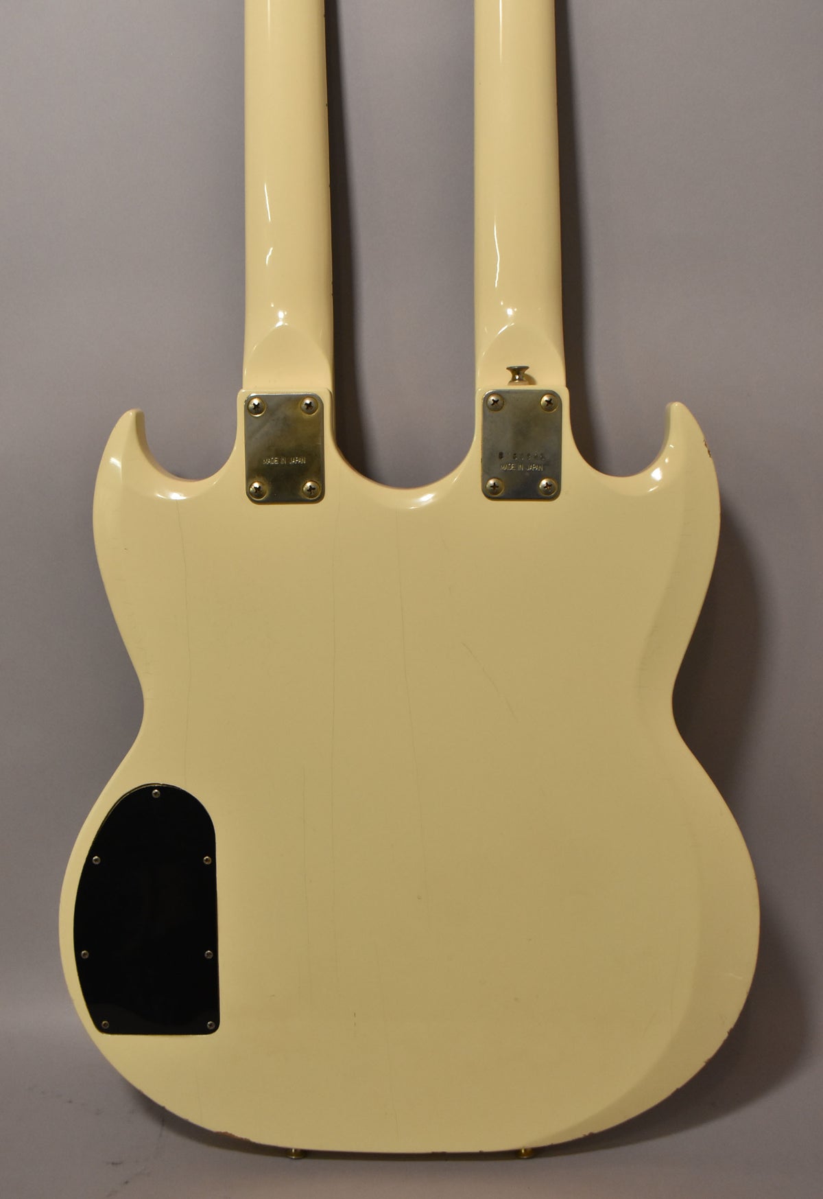 Rear view of a cream double-neck guitar, showing two neck plates, a black control cavity cover, and light finish checking.