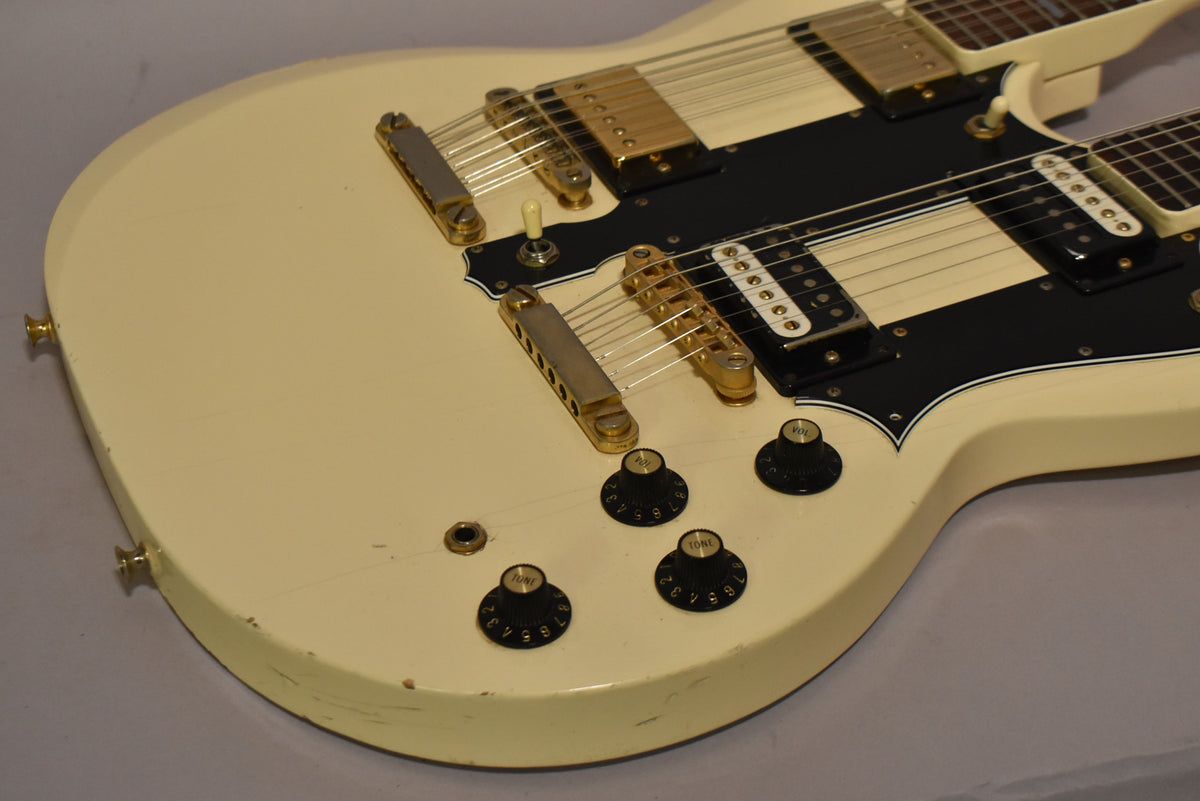 Close-up of the cream-colored double-neck guitar body, showing gold hardware, black pickguard, four black control knobs, and some wear.
