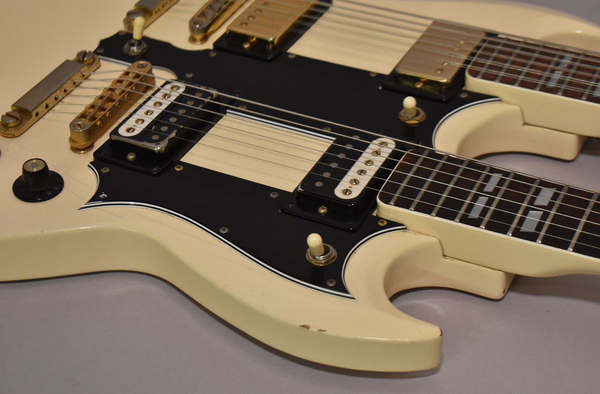 Close-up of the cream double-neck guitar body and black pickguard, showing gold hardware, white humbucker pickups, and minor chips.