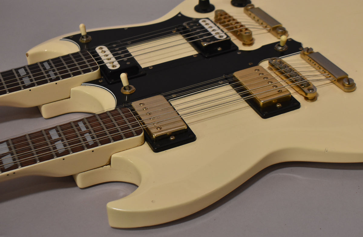 The white double-neck guitar's body is angled from the lower right, revealing two black pickguards, gold hardware, and block inlays.