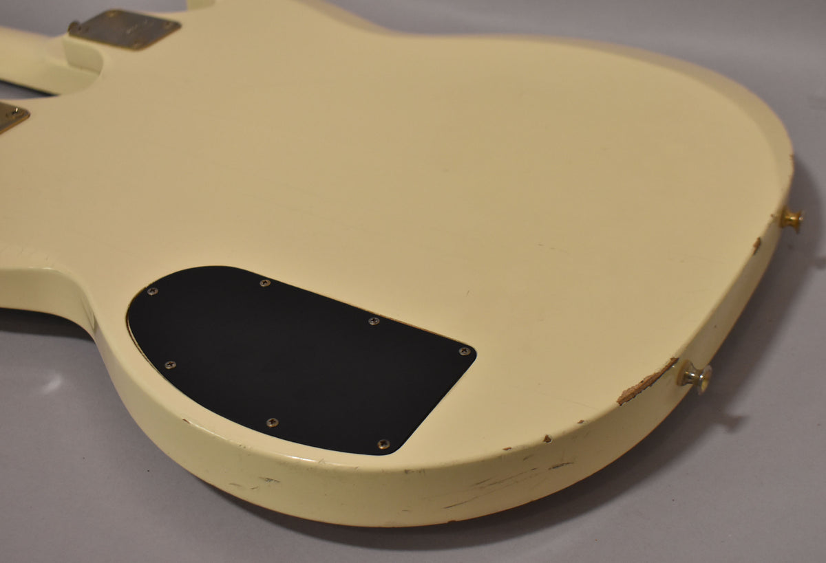 The back of a white double-neck guitar, angled down, showing a black control cavity cover and significant paint chips on the lower edge.