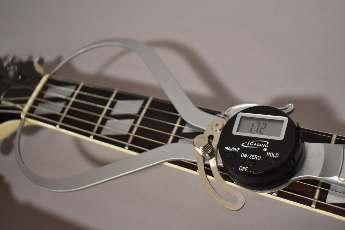 A digital caliper measures the string height at the 12th fret of the guitar's dark fretboard with trapezoid inlays.