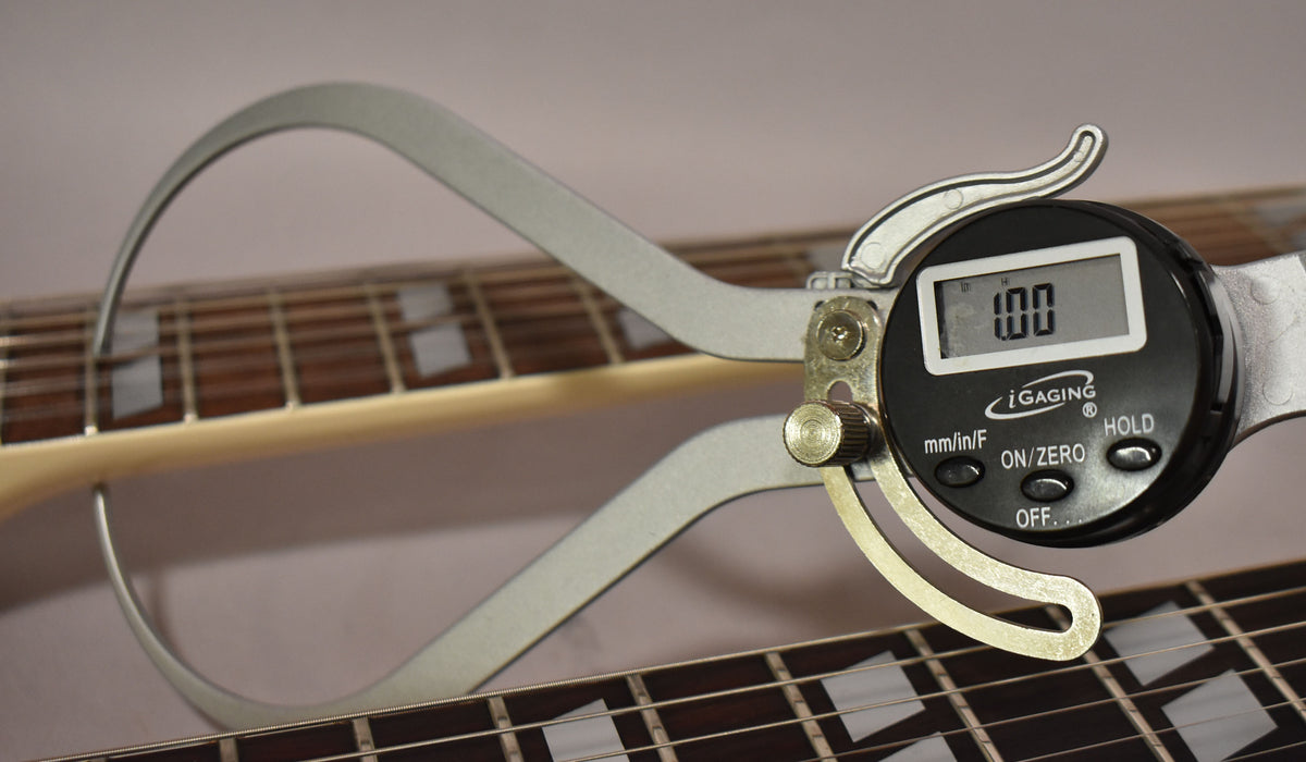 A digital caliper measures the neck thickness of the upper guitar neck, showing "1.00" on its display, with the lower neck visible below.