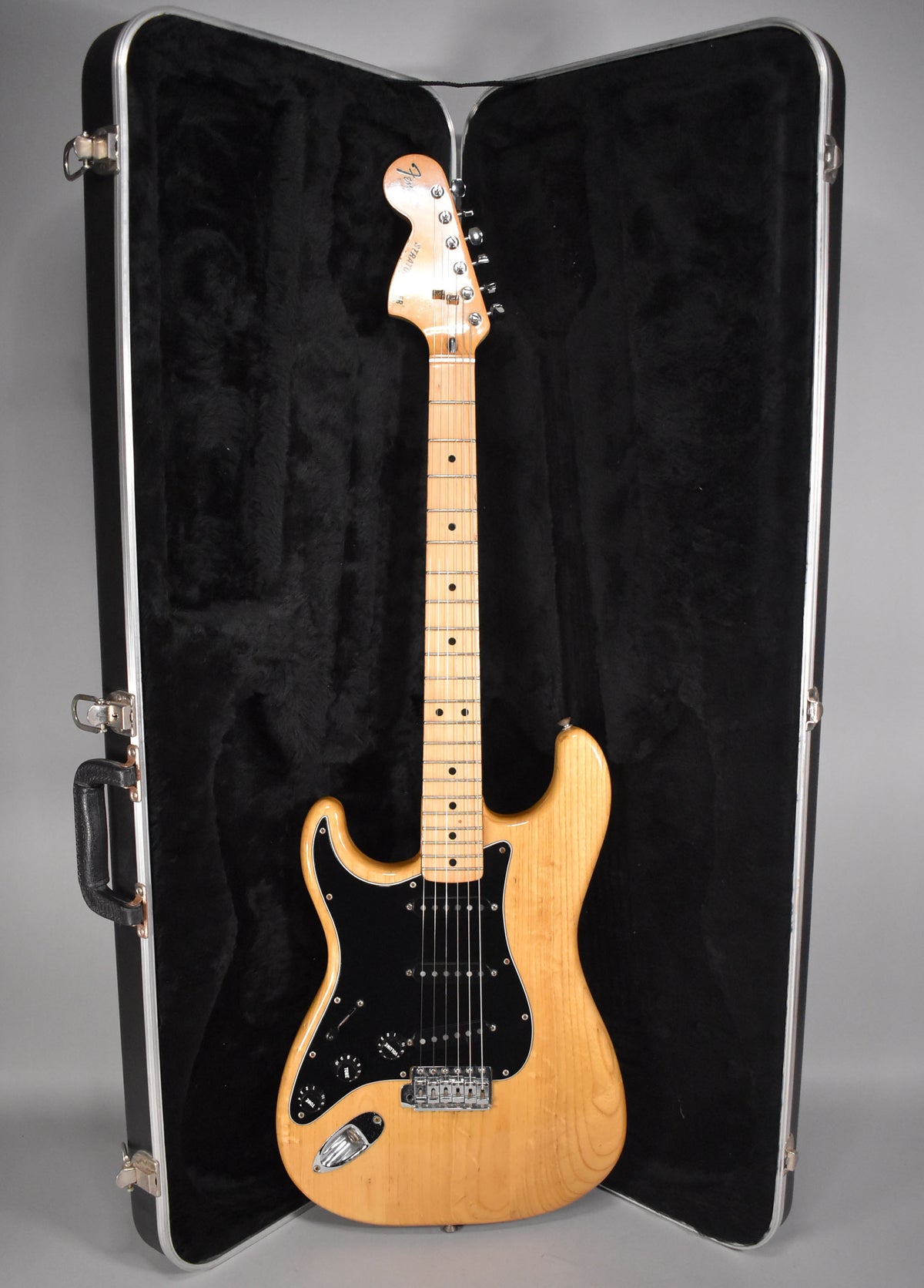 Full-length view of a natural finish Stratocaster with a black pickguard, maple neck, and chrome hardware, in an open hard case.