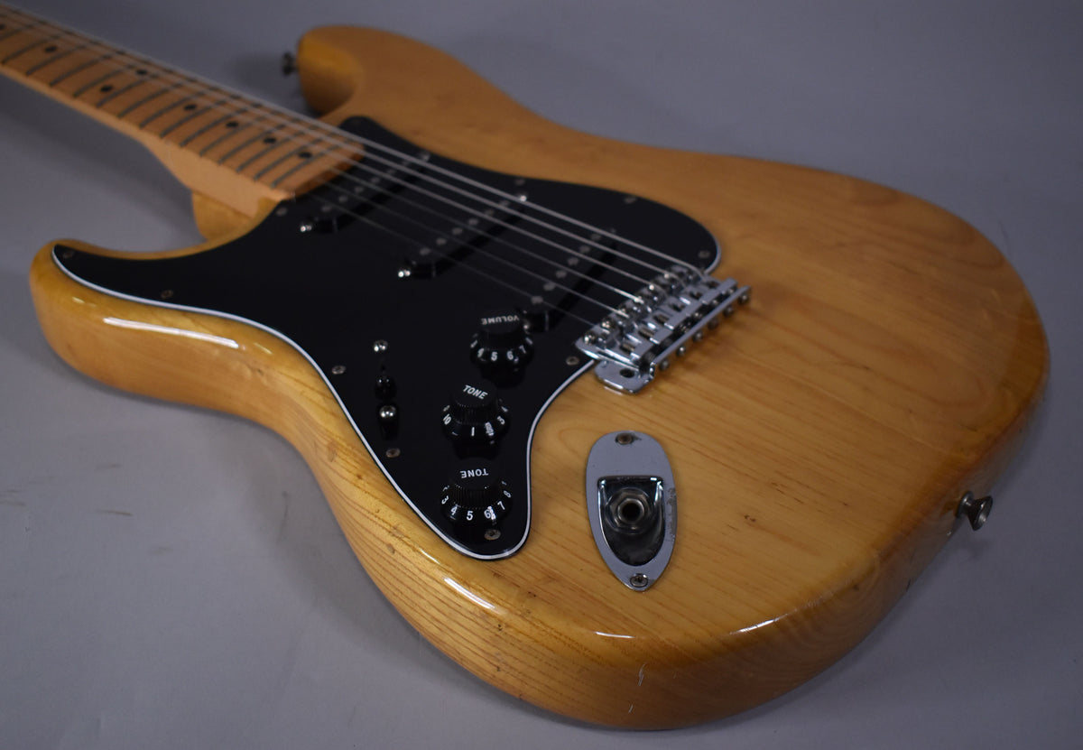Left-handed natural finish electric guitar body with black pickguard, three black knobs, chrome bridge, and visible wear on the lower bout.