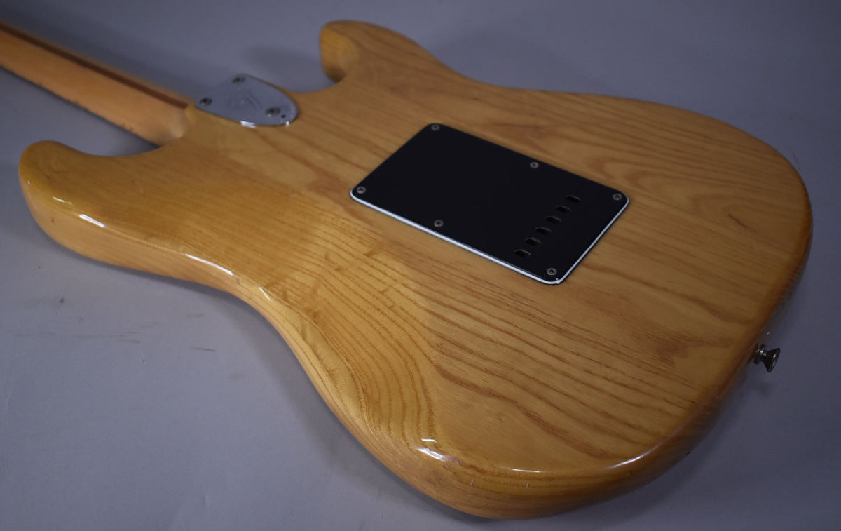 The back of a natural finish Stratocaster, showing wood grain, a black tremolo cover, and a chrome neck plate.