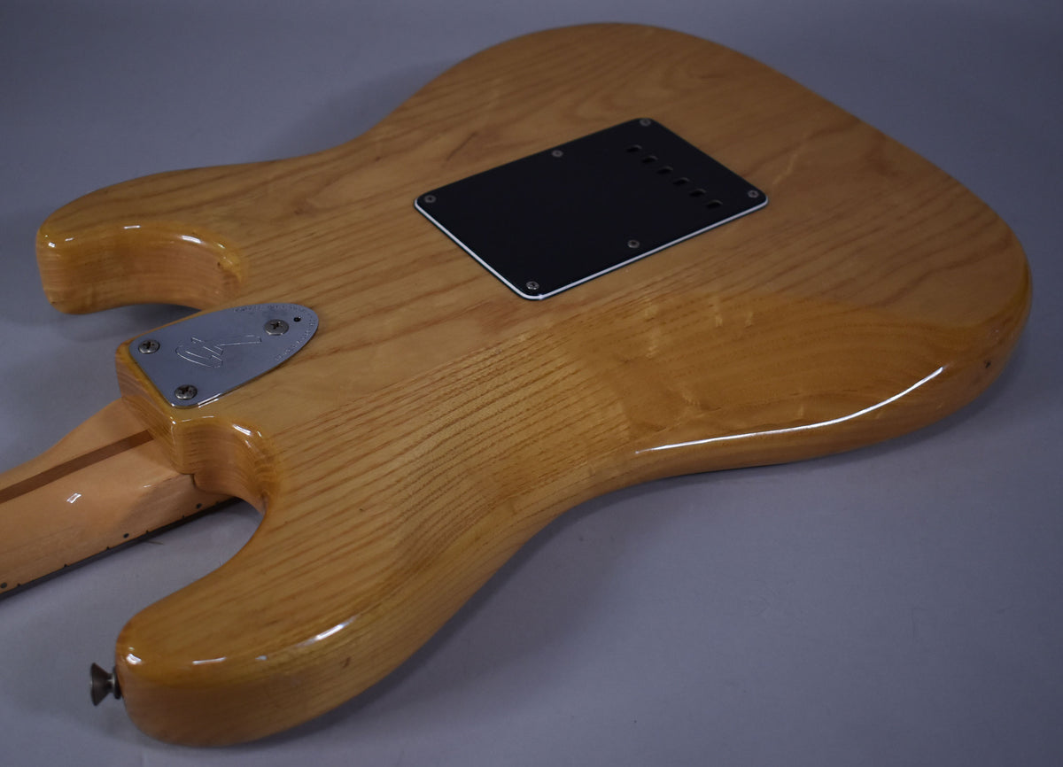 The back of a natural finish Stratocaster, showing the neck plate, black tremolo cover, and light wear on the body edges.