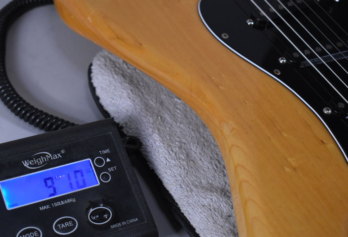 The natural finish body of the guitar rests on a scale displaying 9.10 lbs, with a black pickguard and pickups visible.