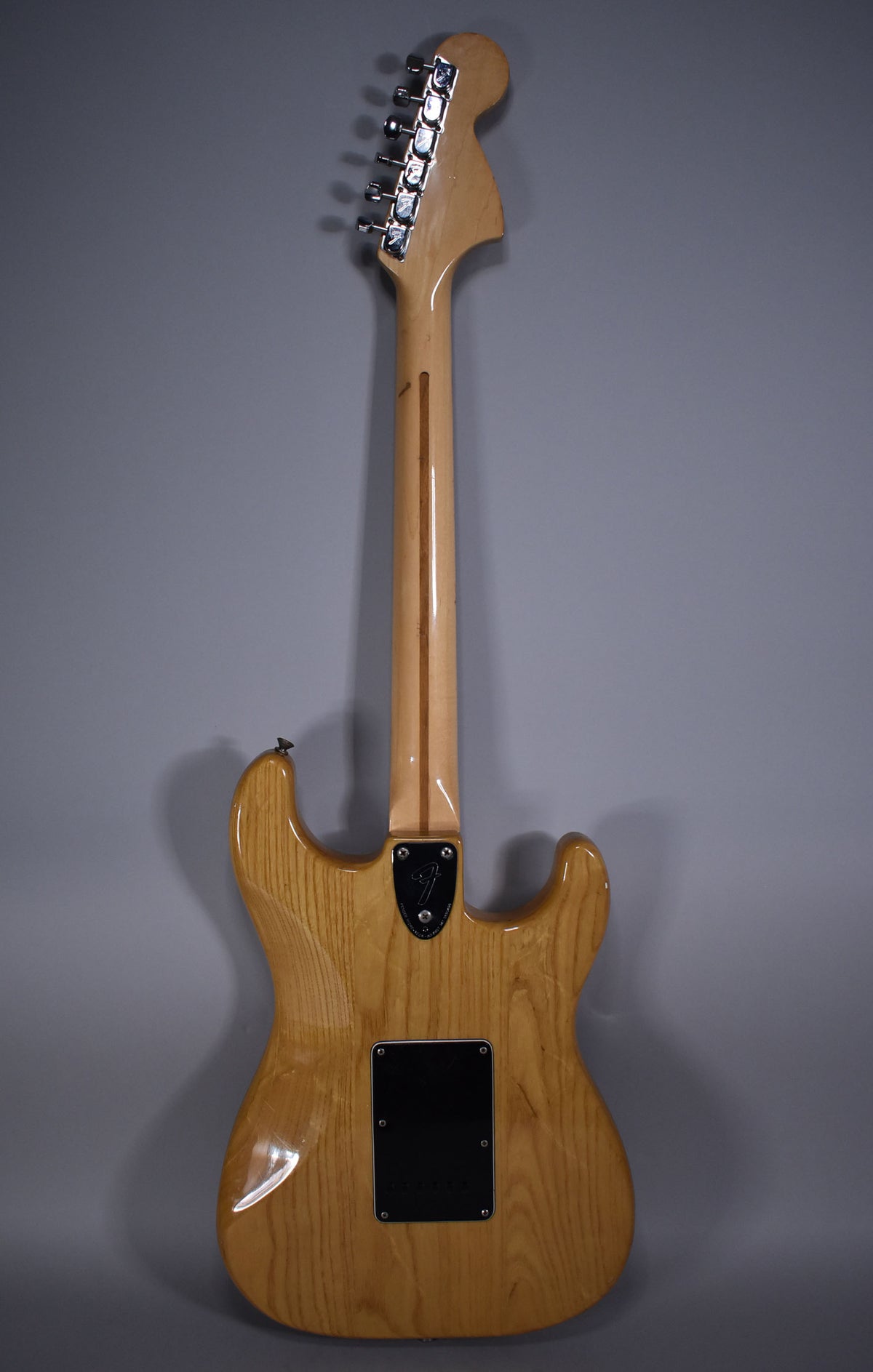 Full rear view of a natural finish Stratocaster, showing the light wood grain, chrome tuners, and black neck plate and tremolo cover.
