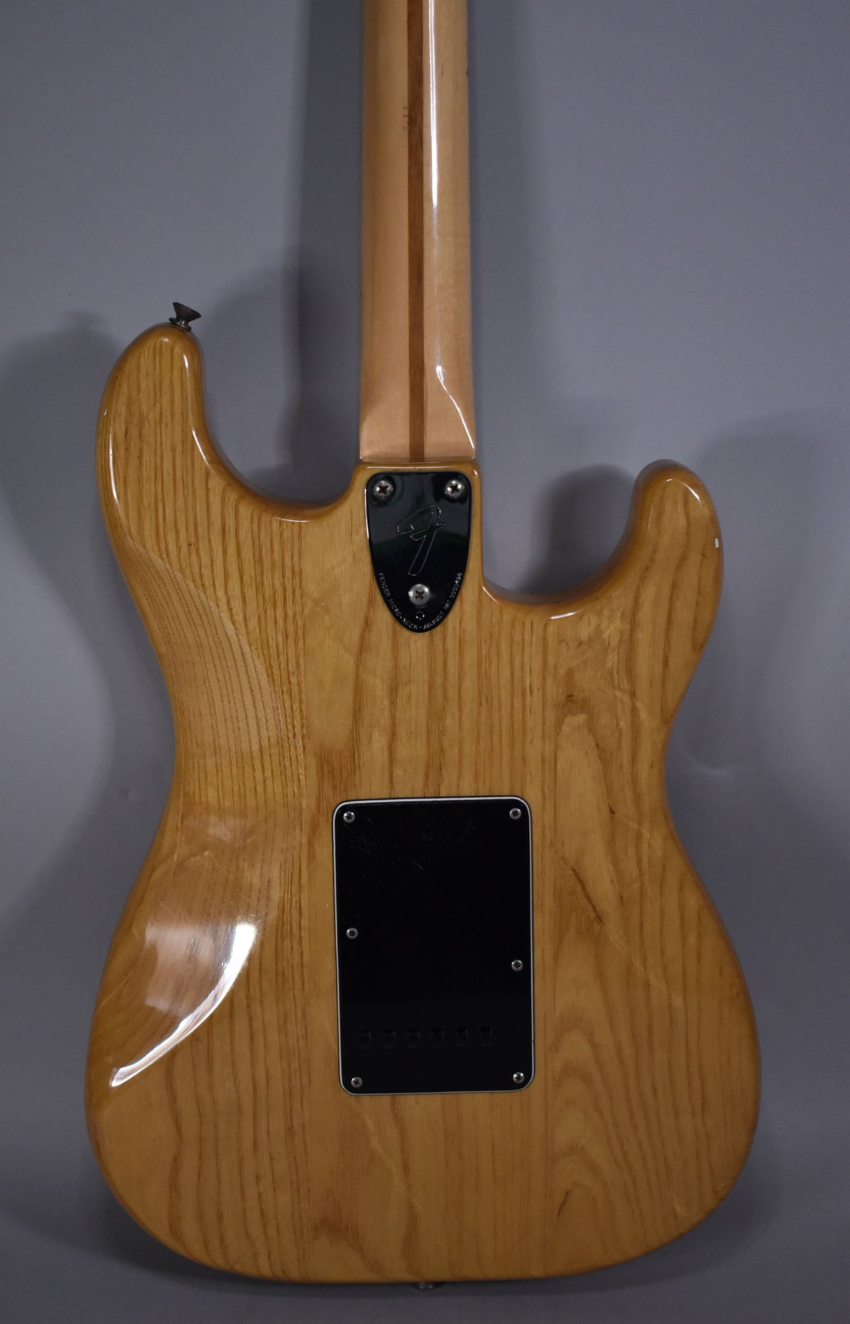 The back of a natural finish Stratocaster, showing the neck plate, black tremolo cover, and light wear on the body.