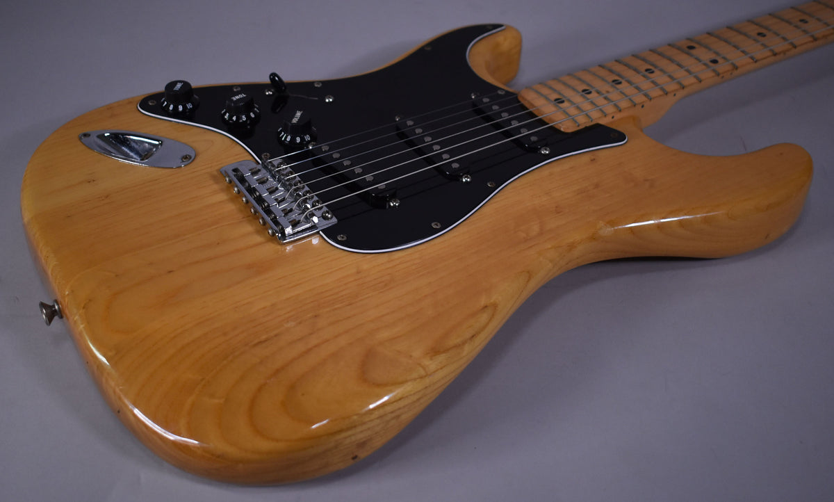 Left-handed natural finish guitar, angled from the lower bout, showing a black pickguard, three black pickups, and chrome hardware.