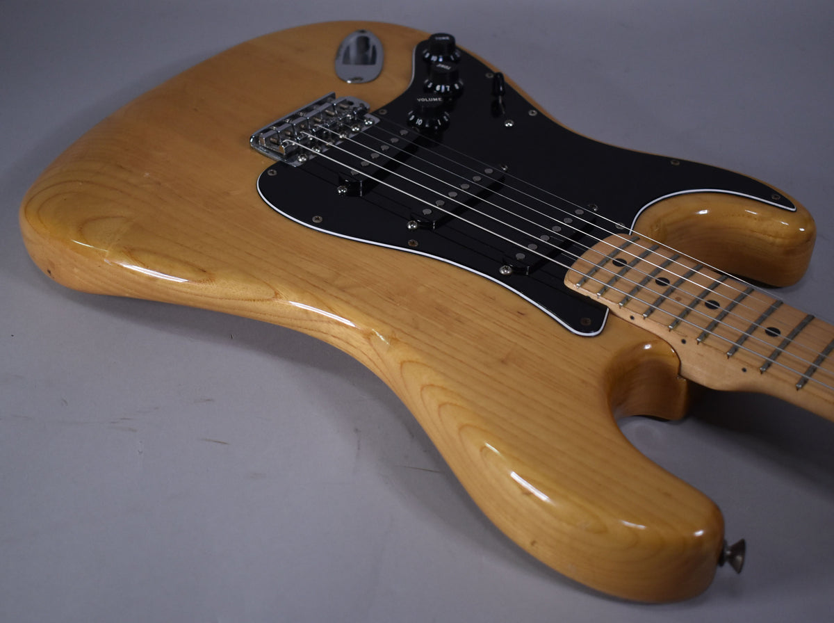 The natural finish guitar body, with a black pickguard and maple fretboard, is angled from the lower right.