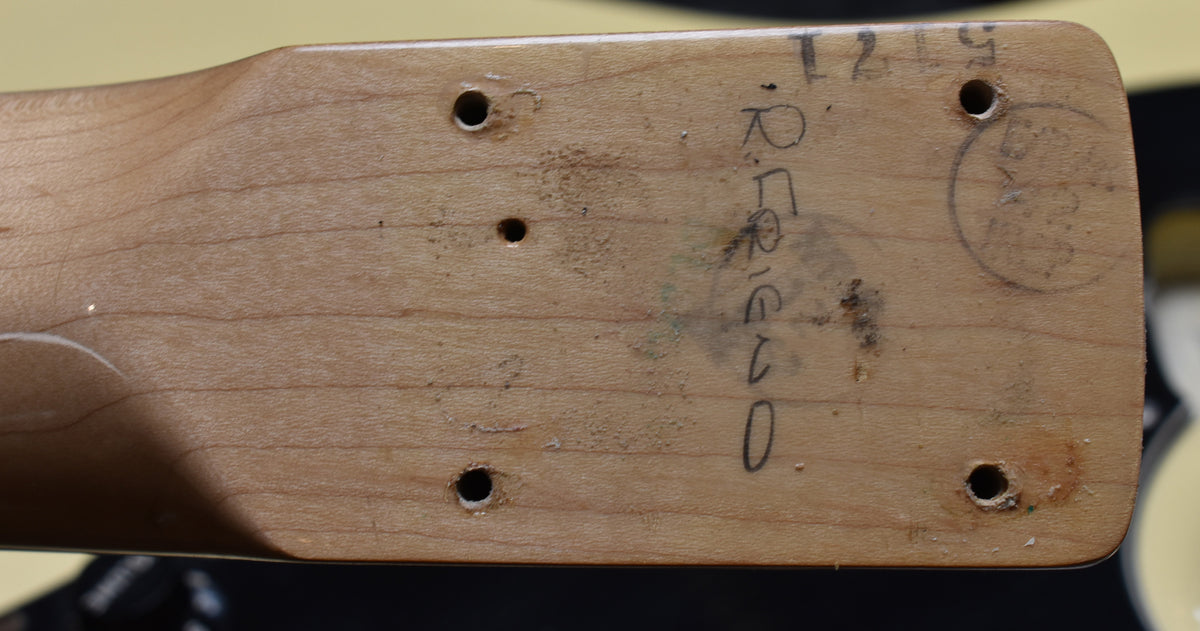 The guitar's neck heel, showing four screw holes, a smaller pilot hole, and various smudges and handwritten markings on the light wood.