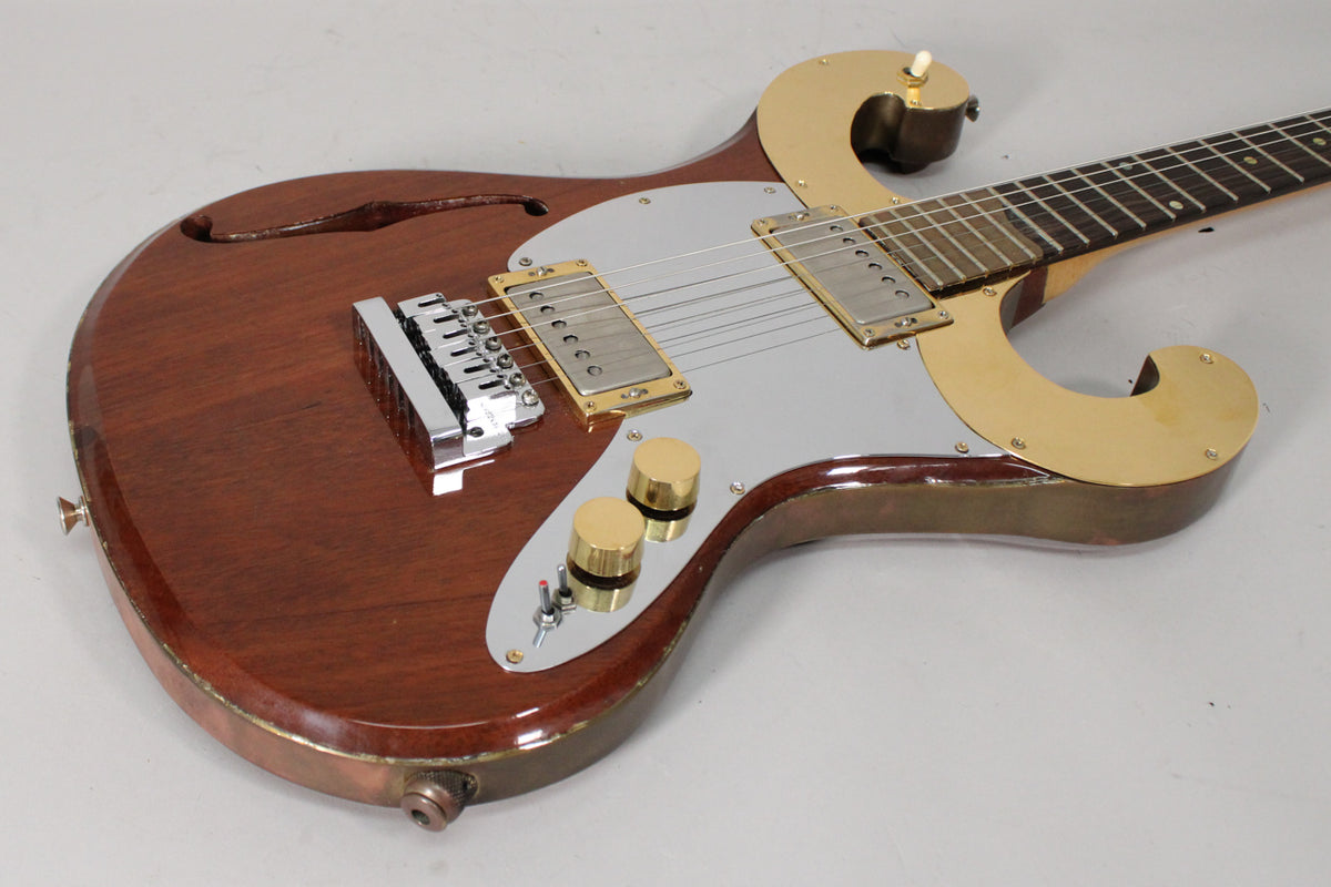 A brown semi-hollow electric guitar with a silver pickguard, gold hardware, and two gold control knobs, viewed from the upper right.