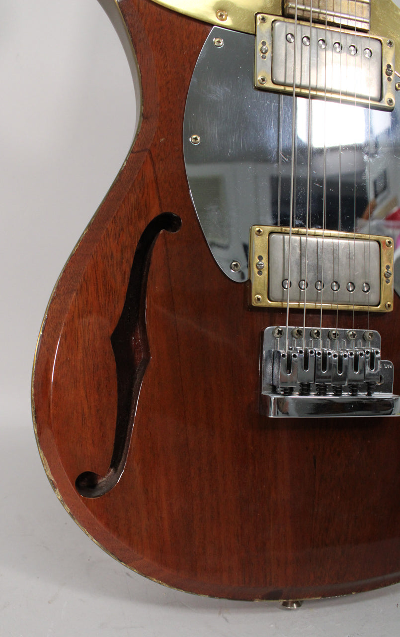 The lower bout of a reddish-brown semi-hollow guitar with an f-hole, chrome pickguard, and two humbuckers with brass surrounds.