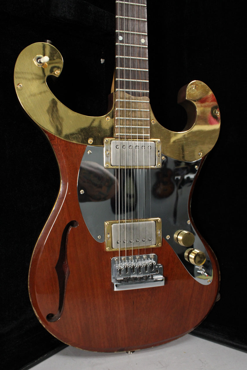 Close-up of a semi-hollowbody electric guitar with a reddish-brown finish, gold hardware, black pickguard, and an f-hole.