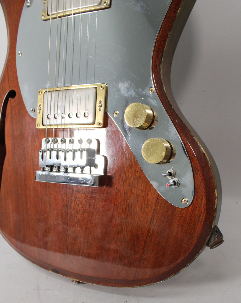 Close-up of the guitar's lower body, showing a worn mahogany finish, scratched chrome pickguard, two brass knobs, and two toggle switches.