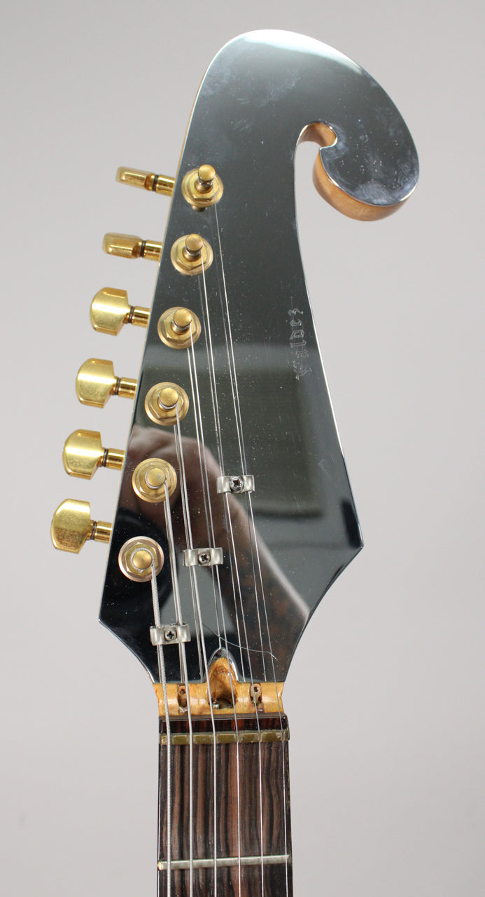 Close-up of the guitar's black mirrored headstock with a curled top, six gold tuners, and a dark wood fretboard.