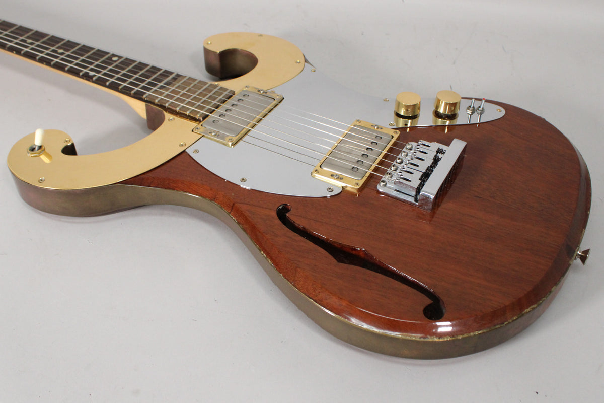 The semi-hollow body electric guitar has a dark wood finish, gold hardware, a silver pickguard, and an f-hole on the lower bout.