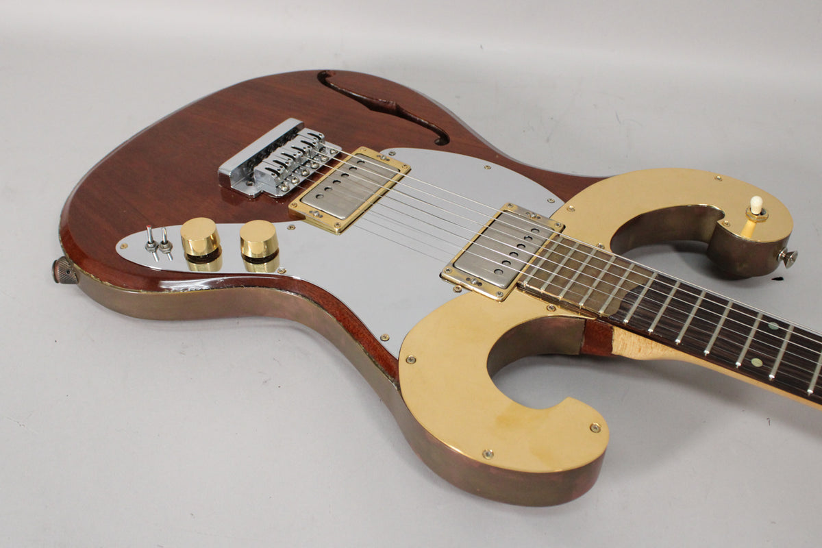The semi-hollow body guitar, in a dark reddish-brown finish, features a silver pickguard, gold hardware, and two gold control knobs.