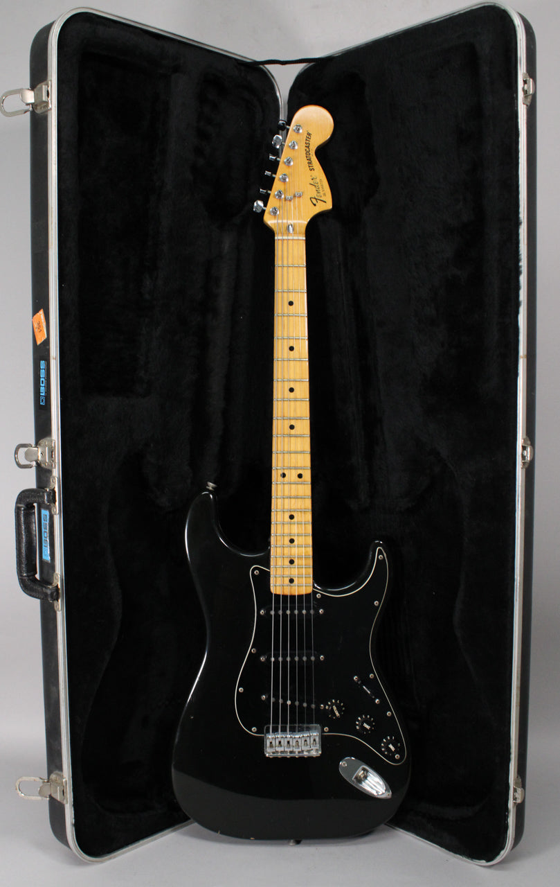 Full-length view of a black Stratocaster with a maple neck and black pickguard, resting in a black plush-lined hard shell case.