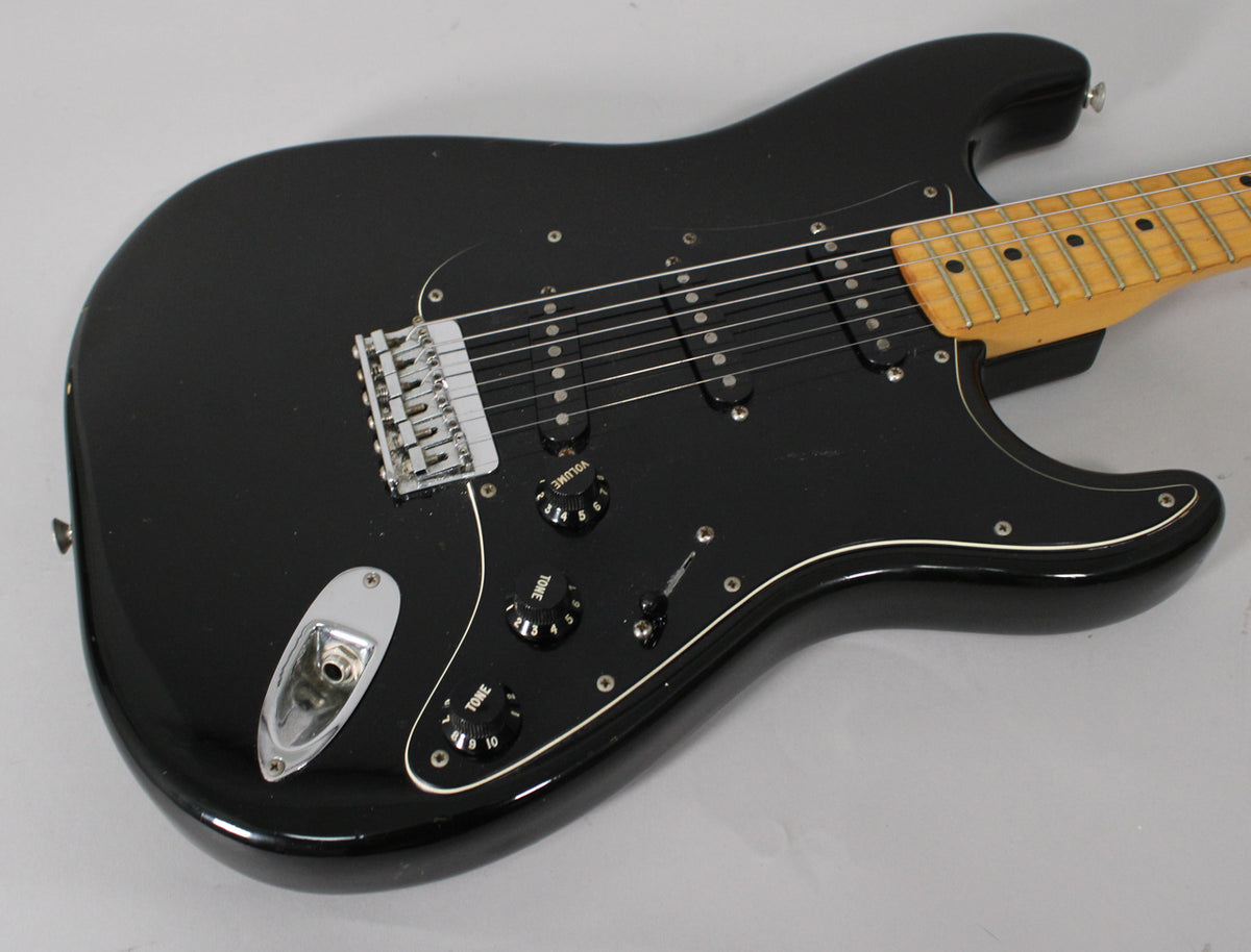 Black Stratocaster body, angled from above, showing the maple neck, black pickguard, chrome hardtail bridge, and three black knobs.