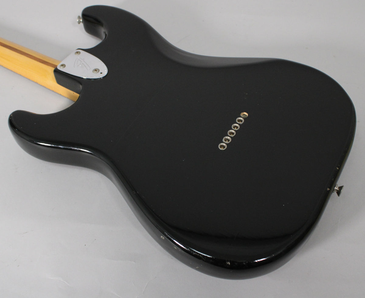 The black guitar body's back is angled, showing six string ferrules, a chrome neck plate, and light wear on the lower edge.