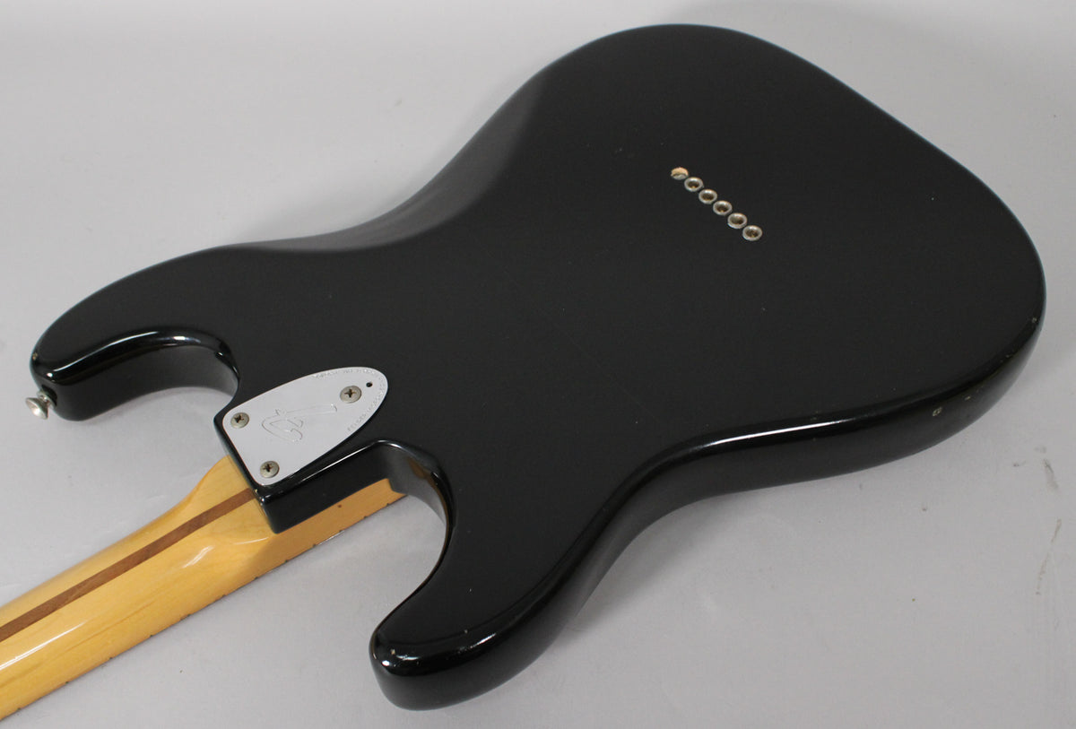 The guitar's glossy black back is angled, showing the neck plate, six string ferrules, and some light wear on the lower bout.