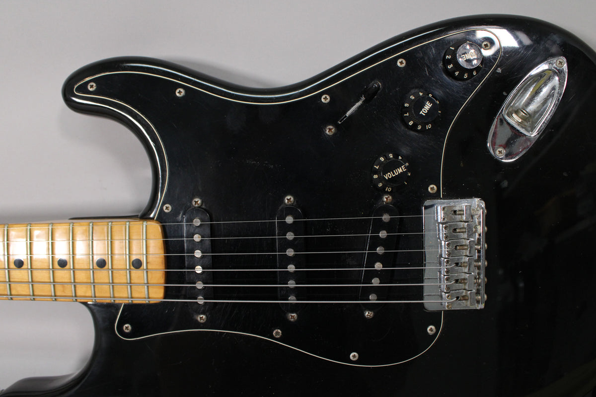Close-up of the black Stratocaster's body, showing the maple neck, three single-coil pickups, chrome hardtail bridge, and worn control knobs.