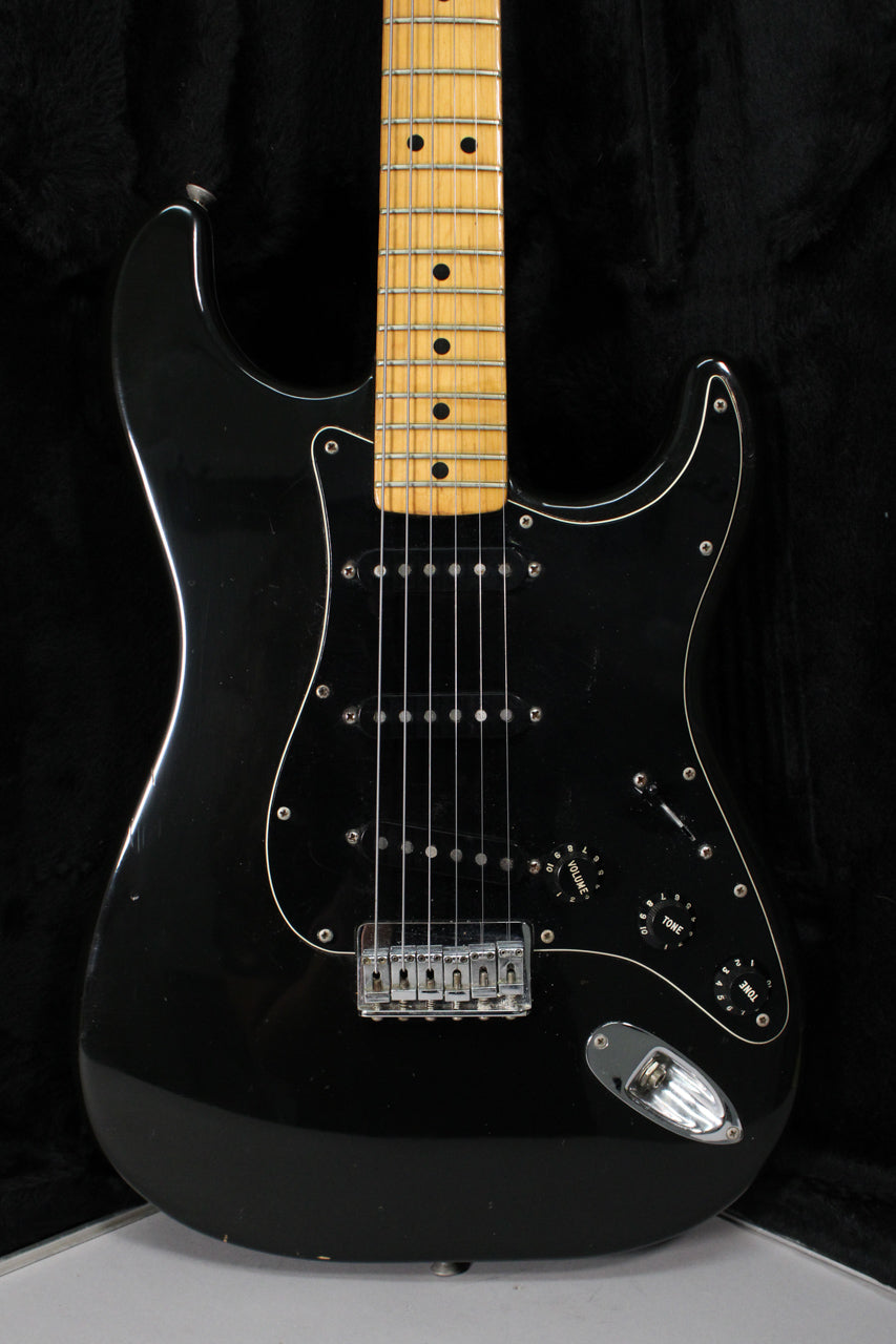 A black Stratocaster, angled from the neck to the lower bout, shows a maple fretboard, black pickguard, and chrome hardtail bridge.