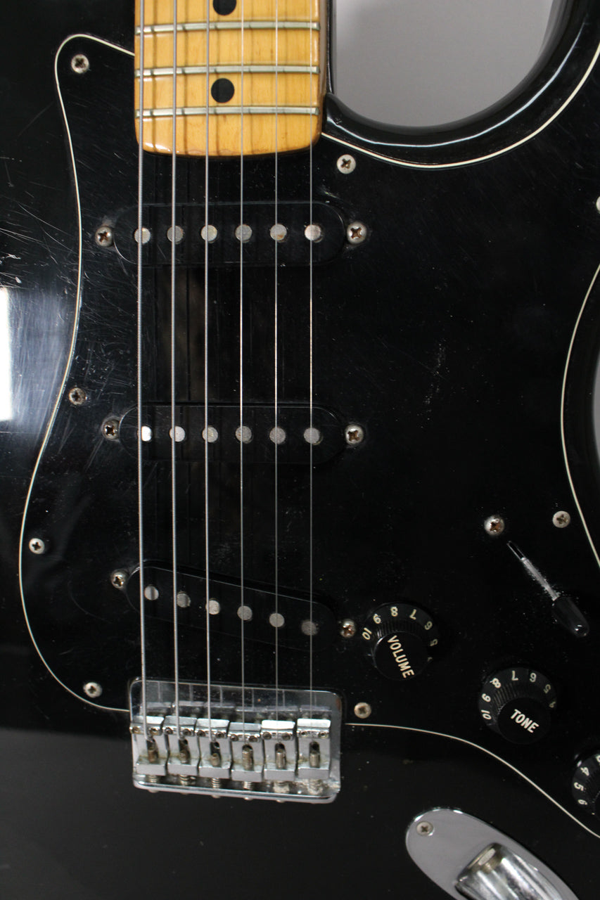 Close-up of a black guitar body with a maple neck, showing three single-coil pickups, chrome hardtail bridge, and wear on the pickguard.