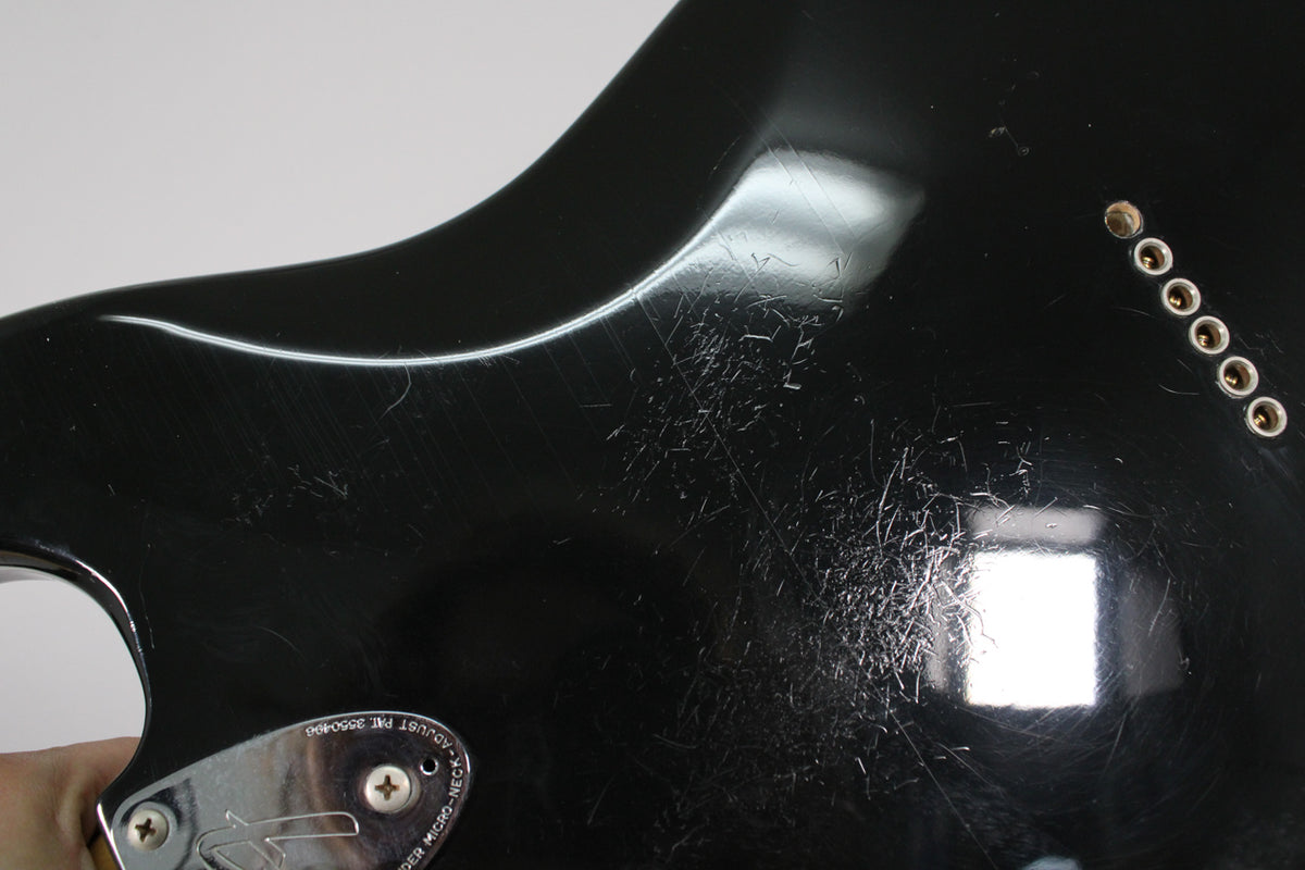 Close-up of the black guitar body's back, showing heavy surface scratches, a chrome neck plate, and six string-through-body ferrules.