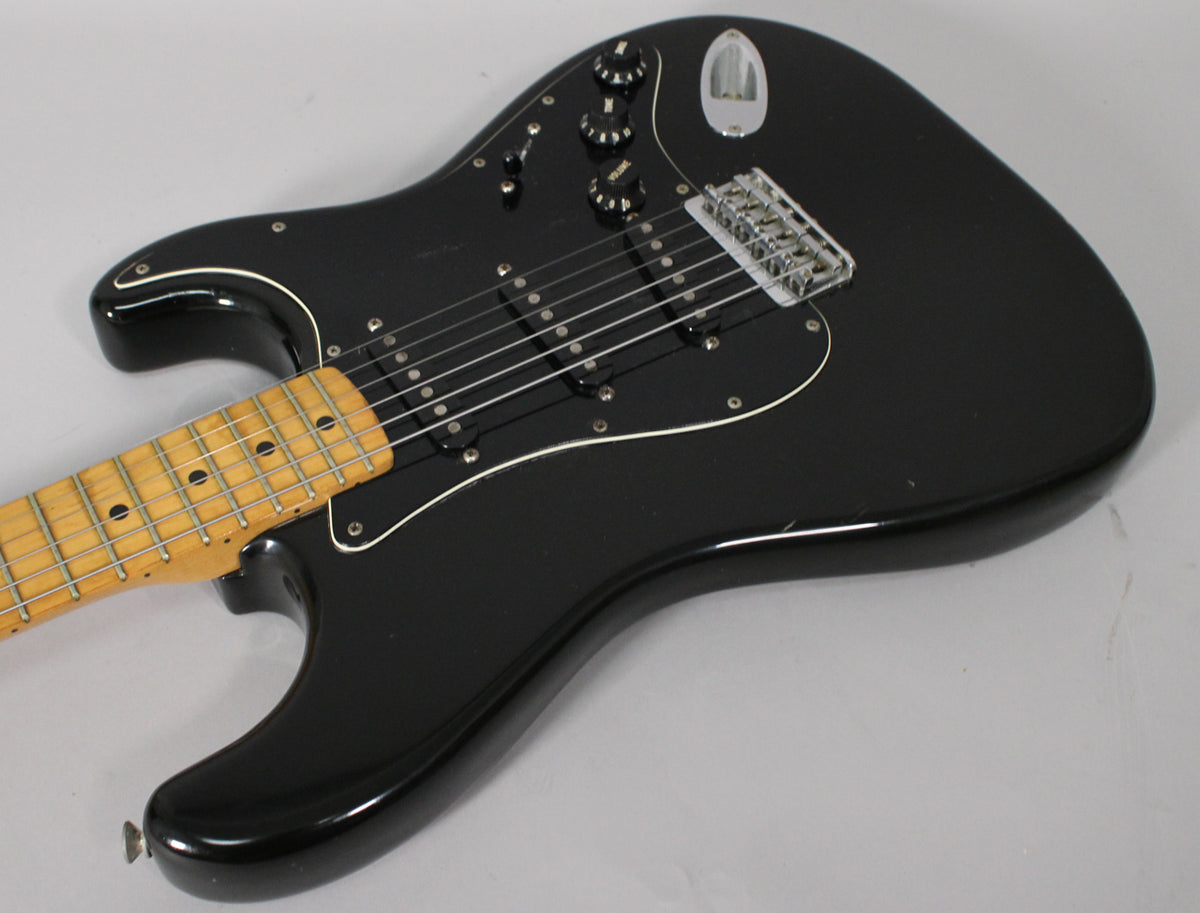 Black electric guitar, angled view of body and maple neck, showing three single-coil pickups, chrome hardtail bridge, and wear on the pickguard.