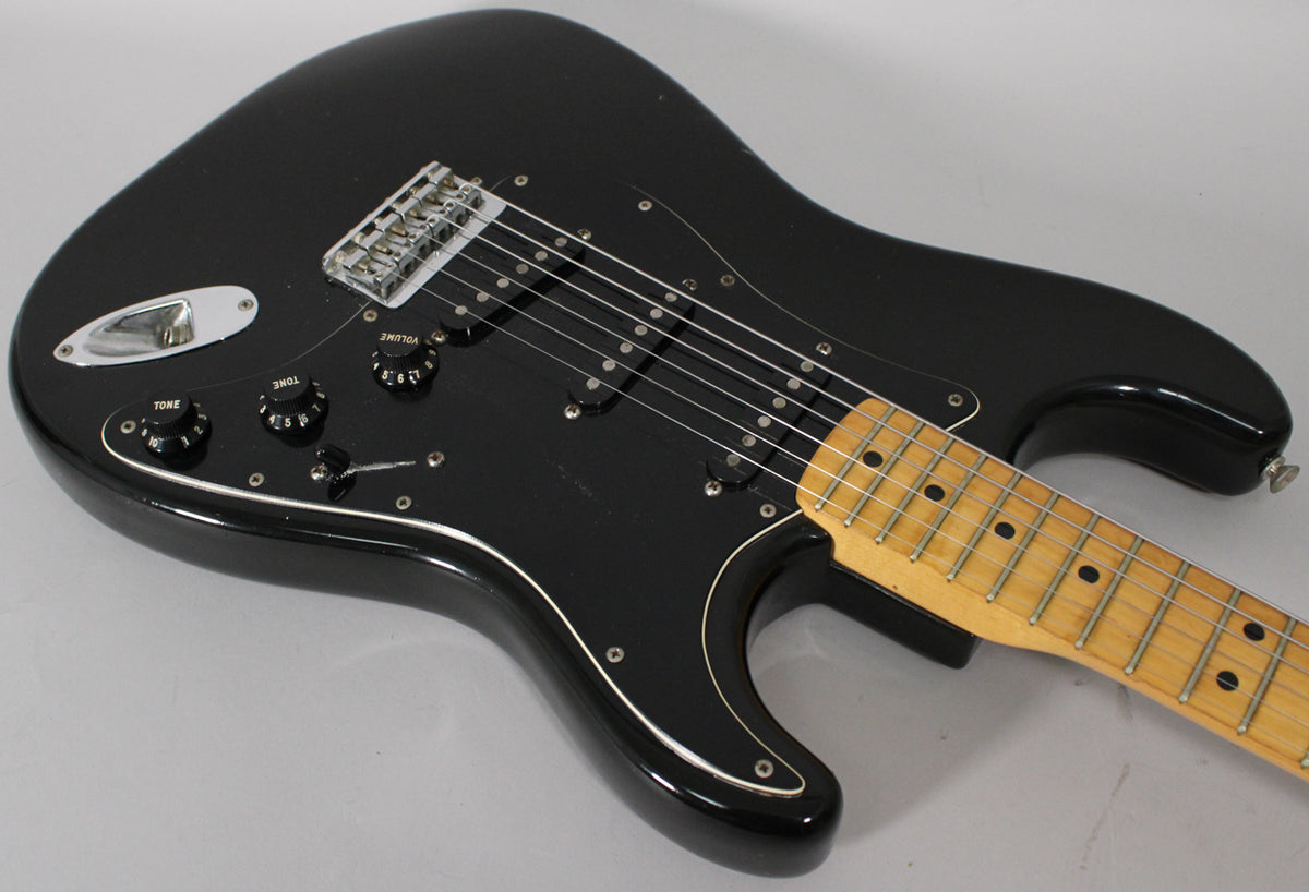 Black Stratocaster body, angled from above, showing the black pickguard, three single-coil pickups, chrome hardtail bridge, and maple fretboard.
