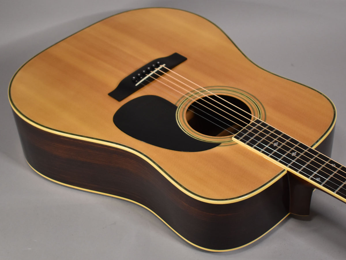 The natural dreadnought acoustic guitar is angled down, showing its light top, dark sides, black pickguard, and part of the fretboard.