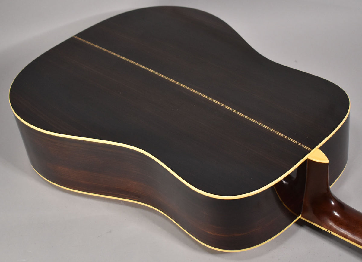 The dark brown acoustic guitar's back is angled, showing a light wood center strip and cream binding along the edges and heel.