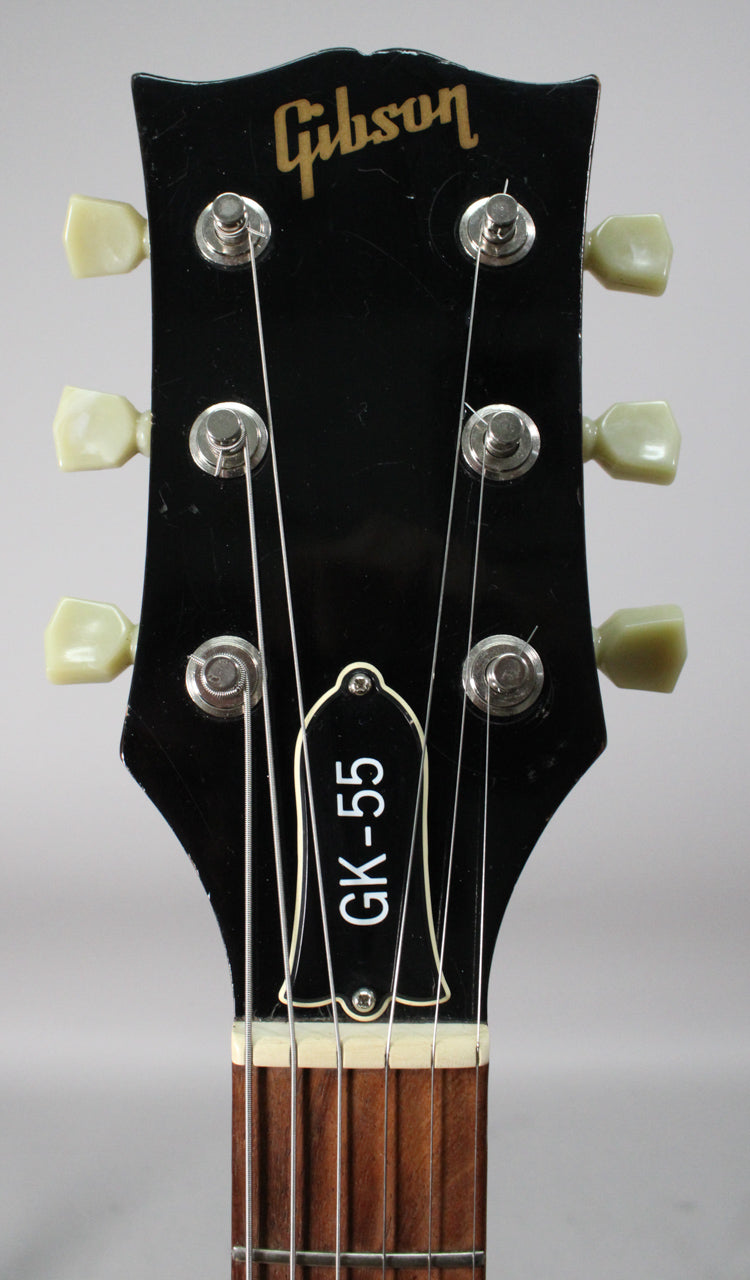 Close-up of the black headstock with "GK-55" on the truss rod cover, six cream tuning pegs, and light wear around the edges.