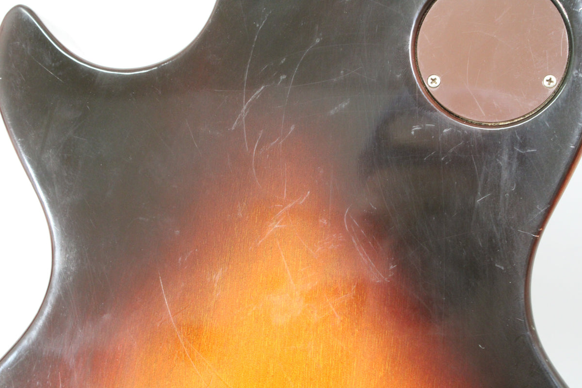 The guitar's tobacco burst back, showing heavy surface scratching and a circular brown control cavity cover with two screws.