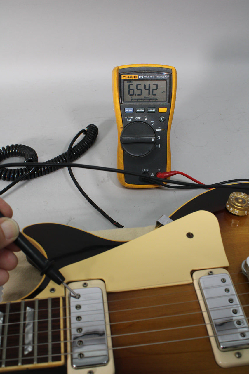 A multimeter reads 6.542 kΩ, connected to the neck pickup of the tobacco burst guitar with cream humbucker rings.