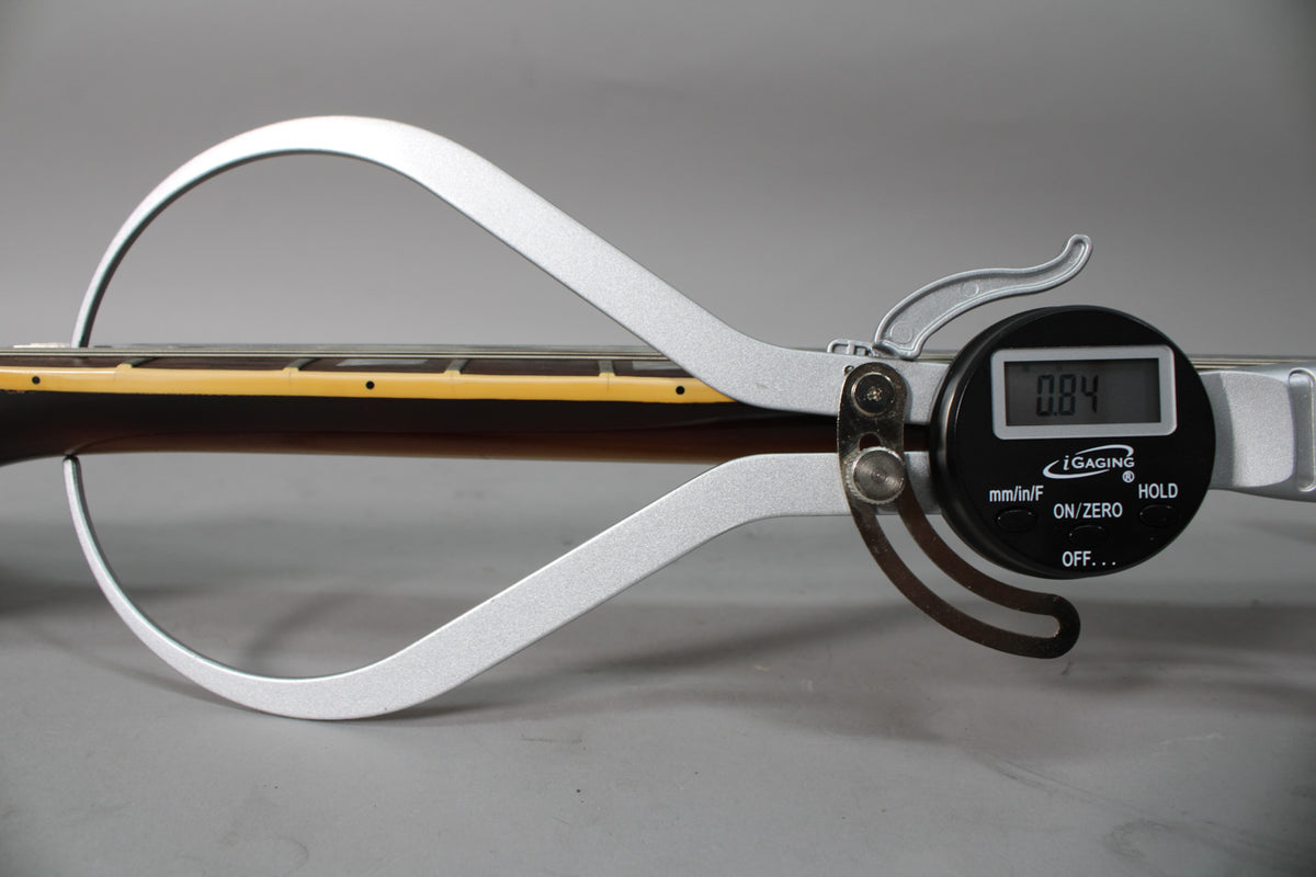 A digital caliper measures the guitar's neck, showing "0.84" on its screen, with the dark brown neck and light fretboard visible.