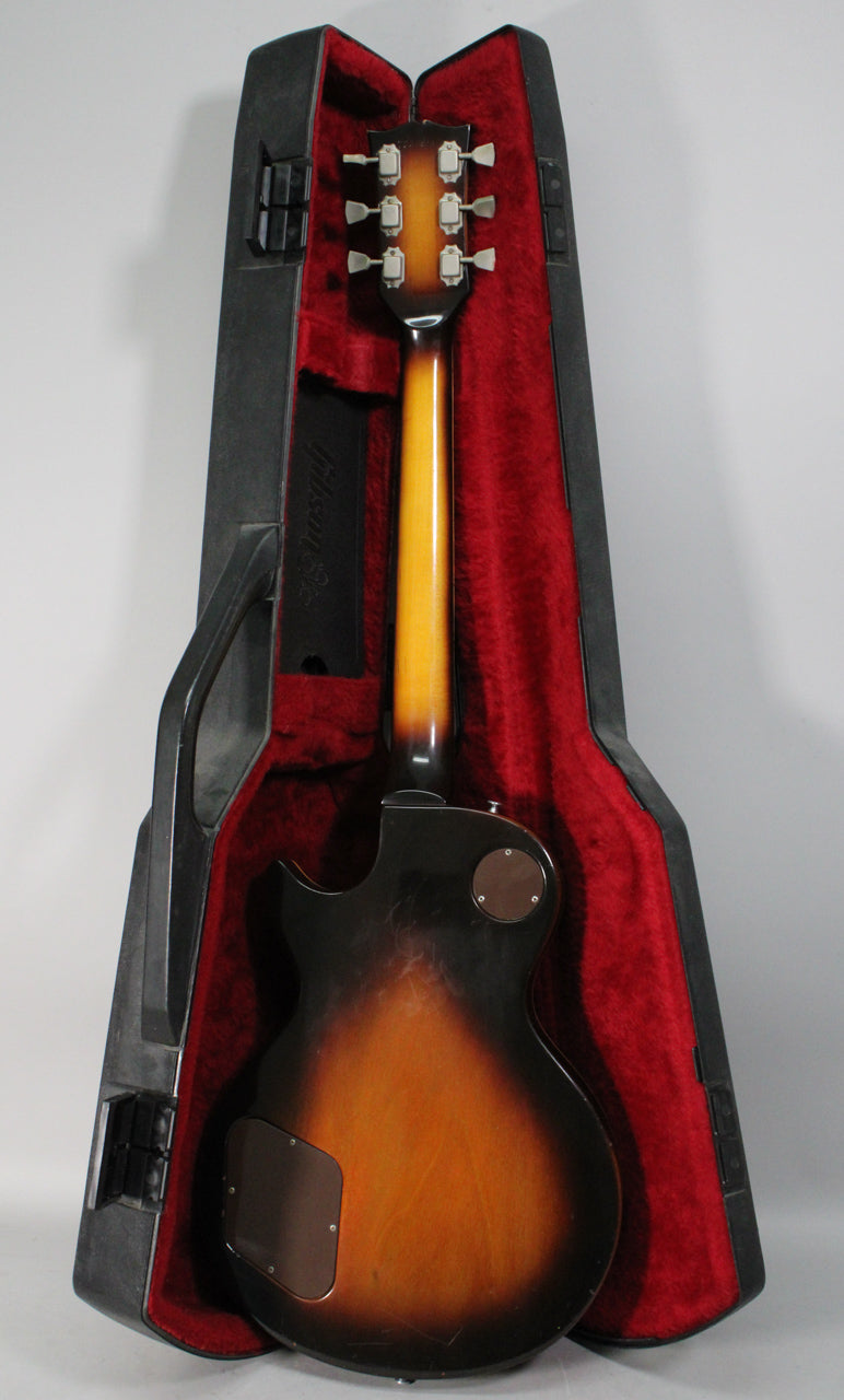 The guitar's tobacco burst back and neck are visible, with a round control plate and rectangular electronics cover, resting in a red-lined case.