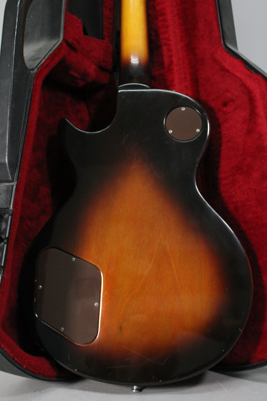 The guitar's tobacco burst back, showing light wear and scratches, rests in a red-lined hard case.