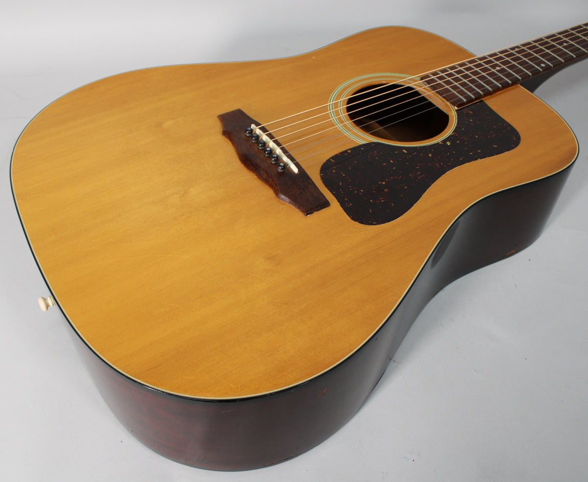 Overhead view of a natural finish dreadnought acoustic guitar, showing the light-colored top, dark sides, pickguard, and bridge.