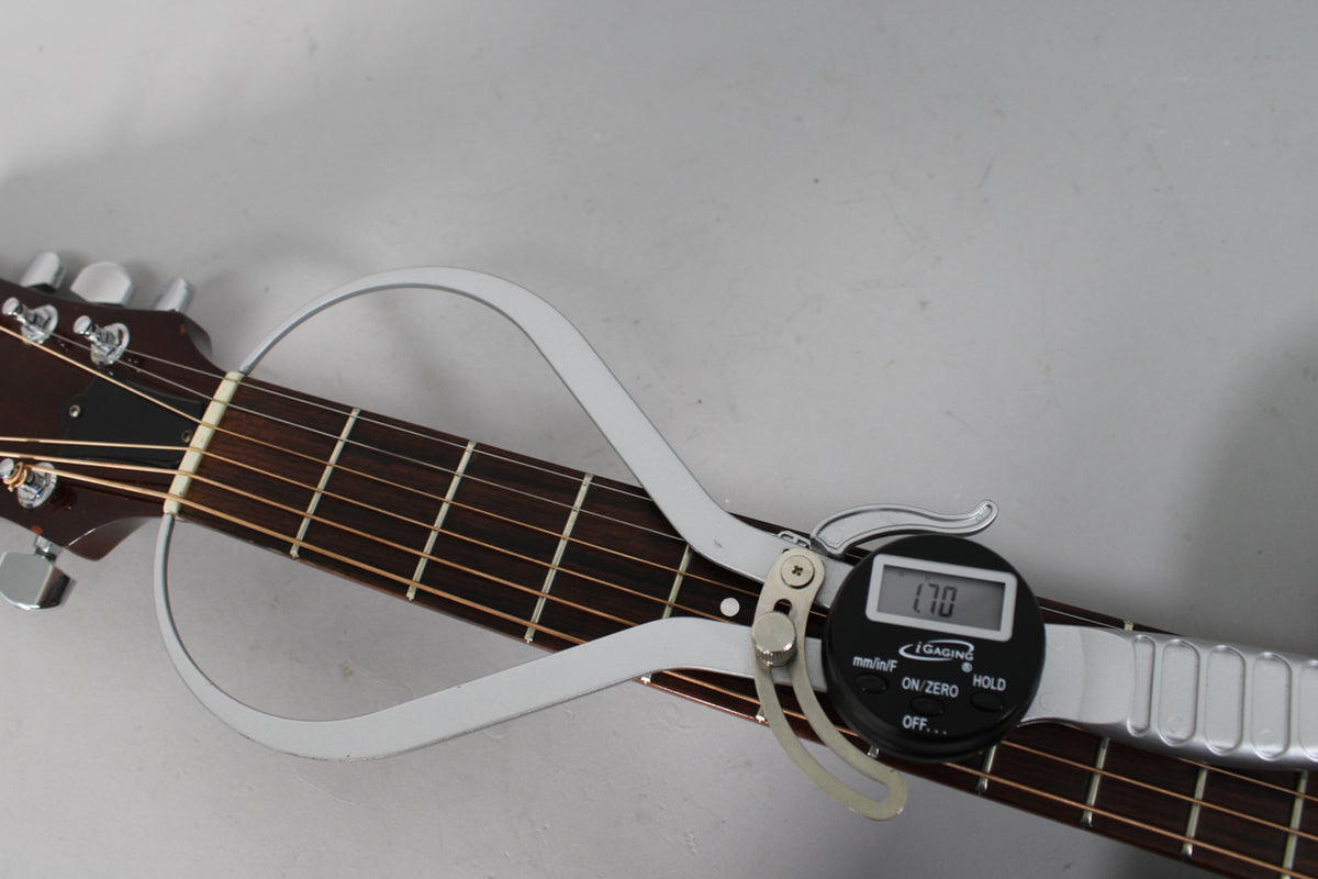 A close-up of the guitar's dark fretboard and headstock, with a silver digital caliper measuring the string height at 1.70mm.