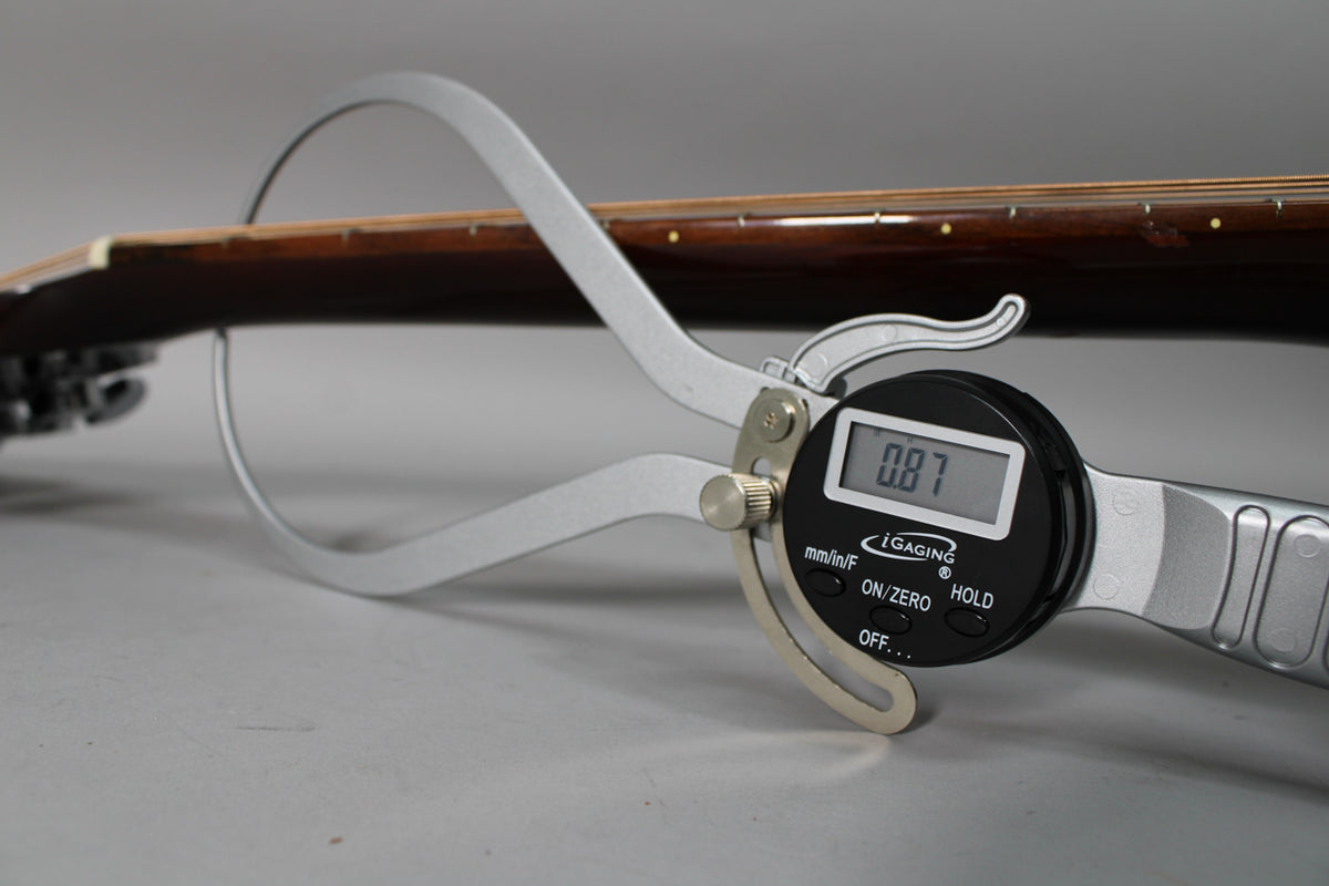 A digital caliper measures the dark brown neck of the guitar, displaying "0.87" on its screen.