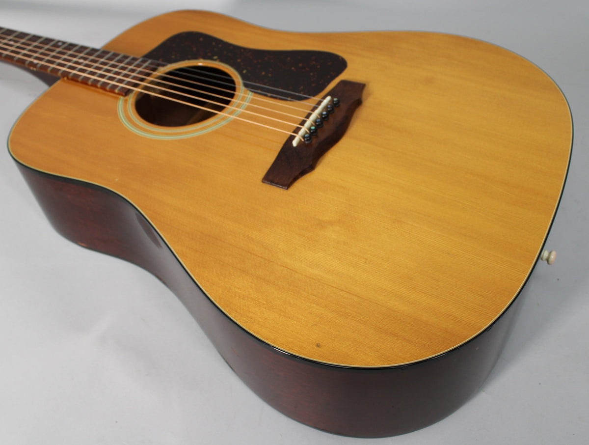 The natural finish acoustic guitar is angled from the upper left, showing the light-colored top and dark sides with some wear.