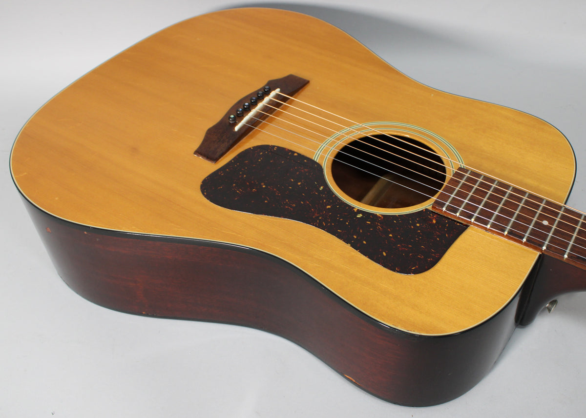 The natural finish acoustic guitar's body is angled, showing the dark pickguard, bridge, soundhole, and lower fretboard, with some wear on the edges.